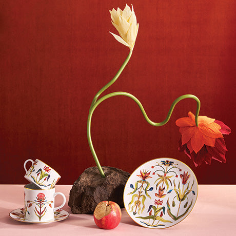 Decorative items including a tulip-shaped vase, teacups, and a plate with floral patterns on a pink surface against a red background.
