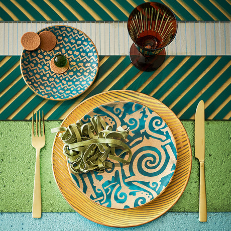 Table setting with patterned plates, pasta, and cutlery on a colorful striped tablecloth.