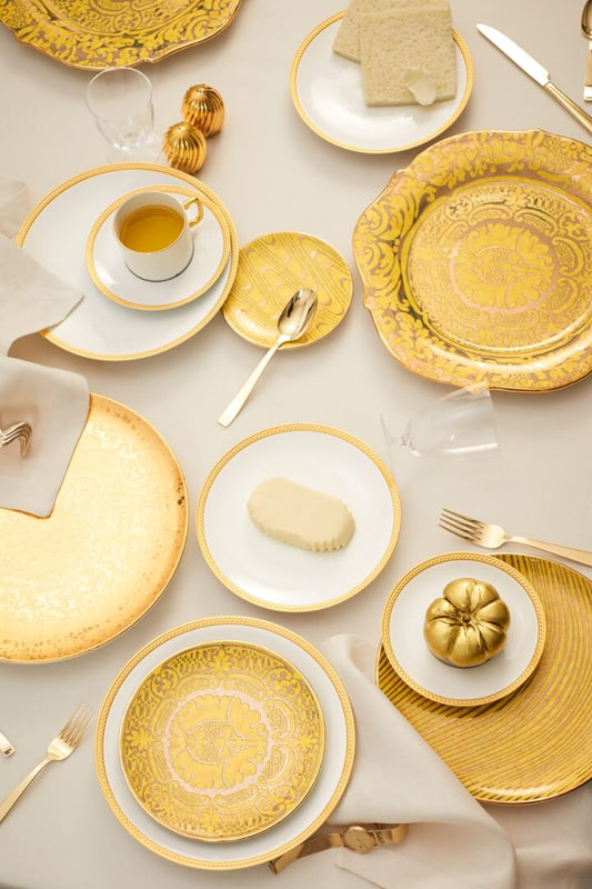 An overhead styled table setting featuring a coordinated collection of gold and white tableware, including patterned plates, charger plates, a teacup and saucer, forks, and napkins on a light neutral background.
