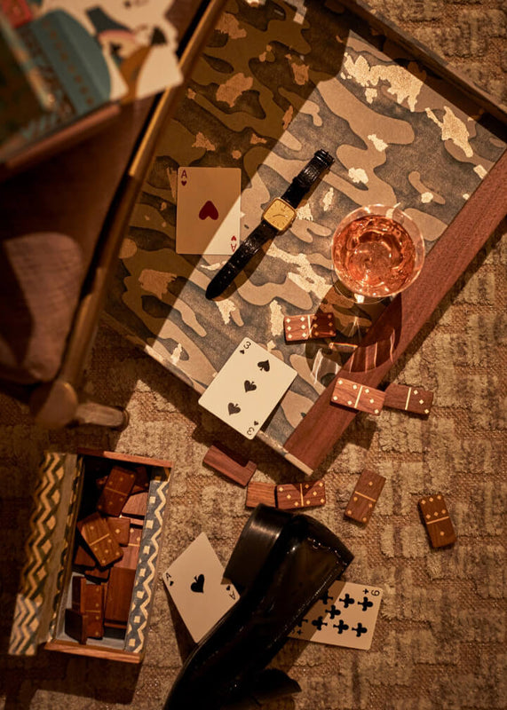 Playing cards, dice, and a watch on a camouflage-patterned surface with a glass of pink liquid.