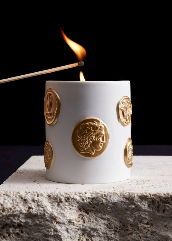 A white cylindrical candle holder decorated with raised gold coin motifs, shown lit with a matchstick and flame against a black background, resting on a textured stone surface.