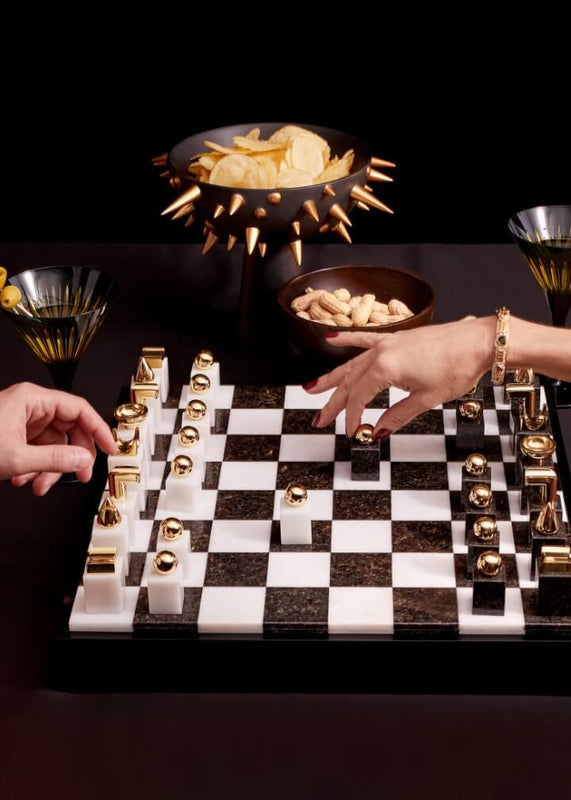 A luxury chess set featuring a black and white marble board with gold-finished chess pieces, styled on a dark table with hands playing with it, snacks in decorative bowls, and gold-rimmed glasses against a black background.