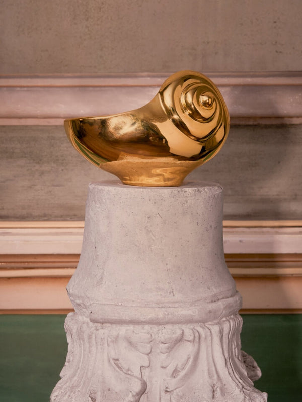 A polished gold metallic sculpture shaped like a spiral shell, displayed on a light gray cylindrical pedestal against a textured beige wall background.